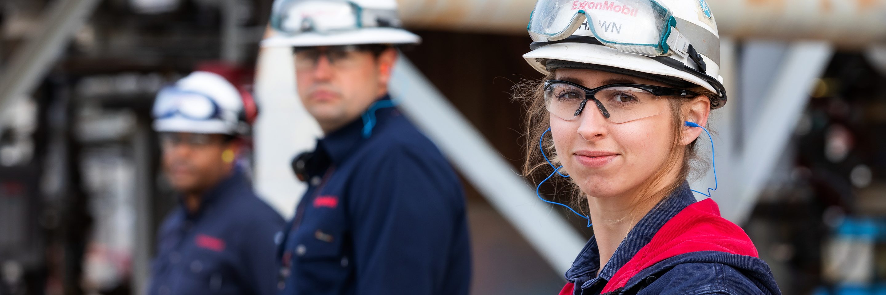 Woman (foreground) and men (background) in hard hats and industrial working glasses, standing on a refinery while they look back at the camera over their left shoulders. 